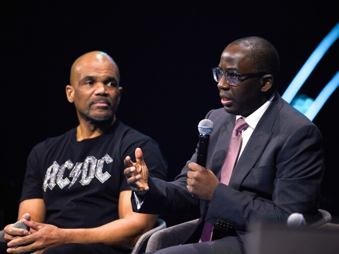 Two men, one with lighter-brown skin tone and another with darker skin tone, sit in chairs on a stage. Both are bald, and the man with darker skin tone is holding a microphone in his left hands as he talks to a crowd that's off camera.