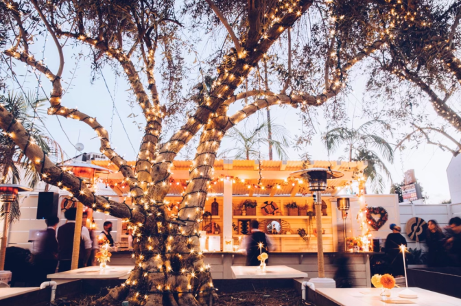 A color image of an outdoor dining setup inside a parking lot. The center focus shows a tall tree wrapped in a string of yellow outdoor lights, with the tops of its branches reaching the top of the image. Below is what looks to be a white outdoor bar area with small groups of people on each side. In front of the bar are metal heat lamps and white tables with centerpieces containing candles and flowers.  