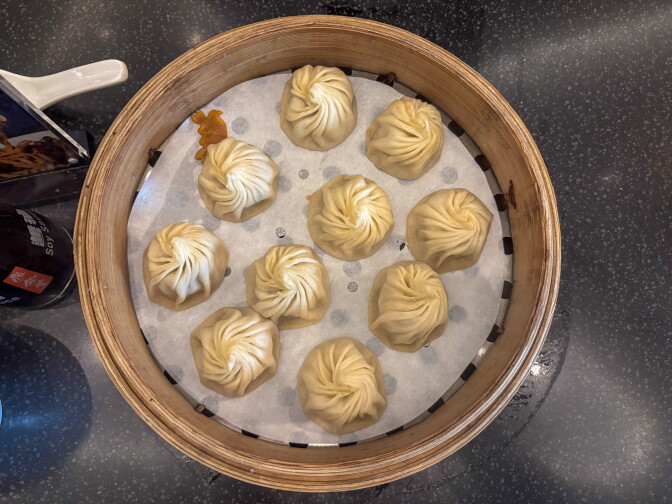 Overhead view of 10 xiao long bao in a round bamboo steamer lined with white paper. Each soup dumpling is pleated into a tight swirl at the top. A small crab-shaped orange garnish sits beside one of the dumplings, and a bottle of soy sauce and a menu holder are at the edge of the dark speckled table.