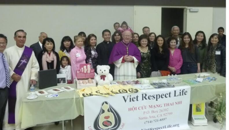 More than a dozen volunteers with a group called Viet Respect Life pose with in front of table with two clergy members.
