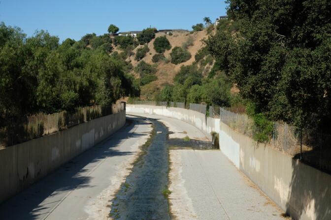 A stream flows in a concrete channel. Chaparral on hills rise behind. 