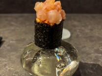 Close-up of a single piece of sushi presented on a decorative glass stand at a restaurant counter. The sushi features pink raw fish or seafood topped with what appears to be uni (sea urchin) or roe, wrapped in dark nori seaweed.