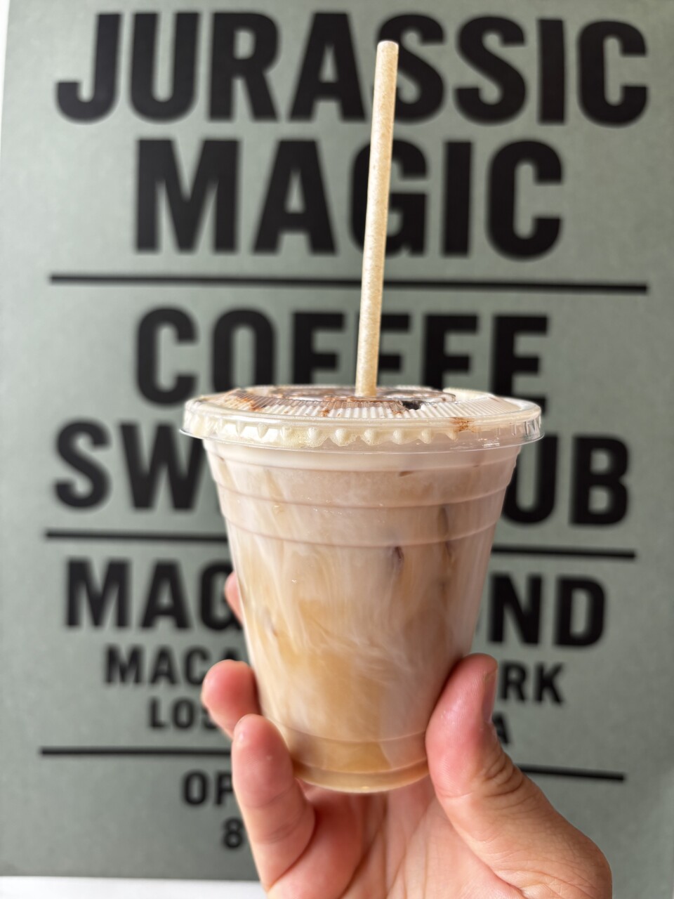 A hand with a light skin tone holds up an iced coffee in a clear plastic cup with a paper straw, in front of a sign that reads “Jurassic Magic Coffee Swap Club” in bold black letters.