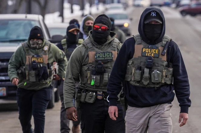 Federal agents, wearing face masks, vests, and some wearing eyewear, walk down a street.