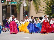 Folkorico dancers wearing skirts in varying colors perform in the street during a parade.