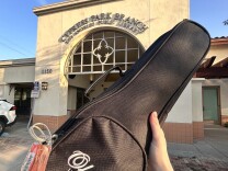 A hand is holding up a black case that says "Ohana Ukuleles" in white font on the front. A large orange laminated tag can be seen on the side, and a tan and brown building is in the background. The building has the text "Cypress Park Branch Los Angeles Public Library" above the entry. 