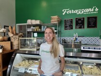 A light-skinned woman stands in front a stainless steel counter with a glass display and trays of rolled pasta behind it. The woman is wearing a white t-shirt. The green wall behind her has a black menu board and a white lettering that reads "Ferrazzani's Pasta & Market."