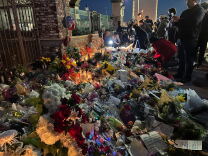 A sea of floral bouquets and candles covers a sidewalk as people and cameras appear in the back.