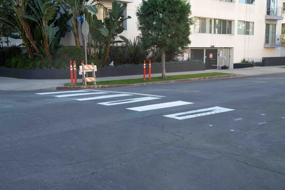 Large rectangles are painted in white on black asphalt. There are bright orange safety bollards and signs laying against a stop sign in the background.