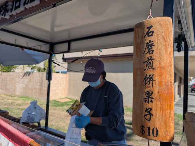 A woman wearing a mask and a baseball cap putting a freshly made crepe into a bag. She is behind a food cart parked on the street.