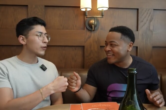 A young Latino man in an oatmeal colored t-shirt wearing glasses fist bumps a young Black man in a black t-shirt while sitting in a restaurant booth 