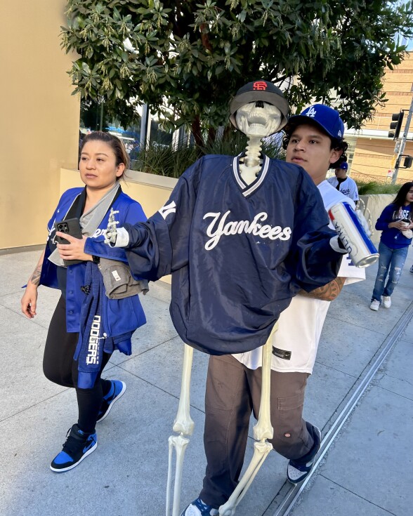 Two people in Dodgers gear  walk with a sketelon dressed in a Yankees pullover and a San Francisco Giants cap