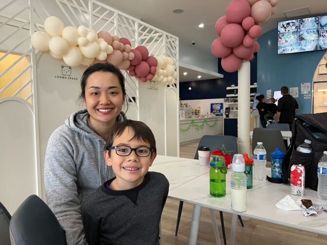 A young male wearing a grey t-shirt and dark glasses smiles. Beside him is a female with medium skin tone and dark hair, wearing a grey hoodie. 