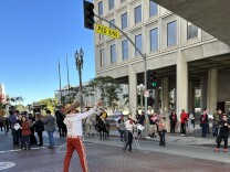 A man wearing a brick red pants and a white shirt uses a lasso in front of a grey building as onlookers look on. 