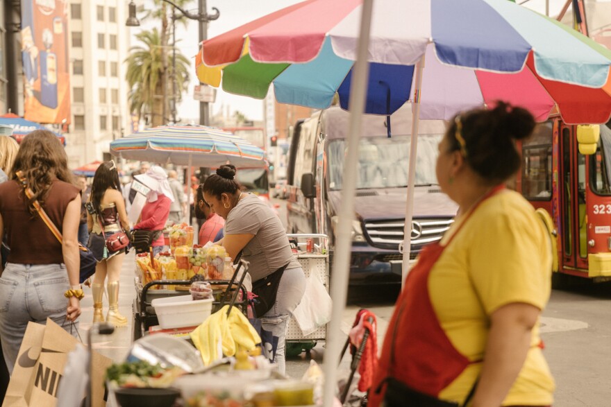 On the sidewalk of a busy city street, a woman at far right in a yellow t-shirt and red apron watches another woman sell food from a street cart under a multi-colored umbrella.