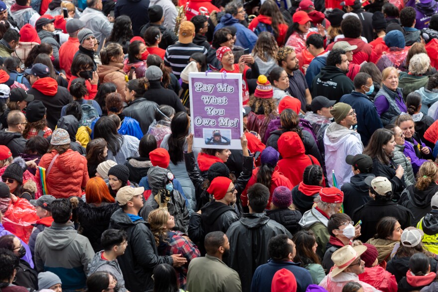 A crowd of people dressed warmly, seen from above. In the midst of the crowd, someone has both hands raised high holding a purple sign up that reads "Pay me what you owe me!"