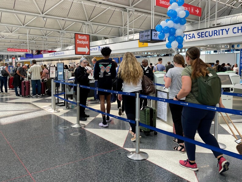 A singe-file line of people wait to check in at an airport terminal. They are corralled by blue ropes. Most are dressed casually. The floor is white and gray.