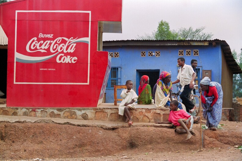 A group of Rwandans waits for transportation near a Coca Cola placard. The photo is from 1994.