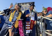 Two young women in graduation regalia walk through a crowd of people gathered in stands.