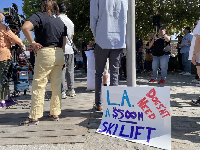 A view from behind of people standing on a sidewalk during a protest. A sign says "LA doesn't need a $500m ski lift."