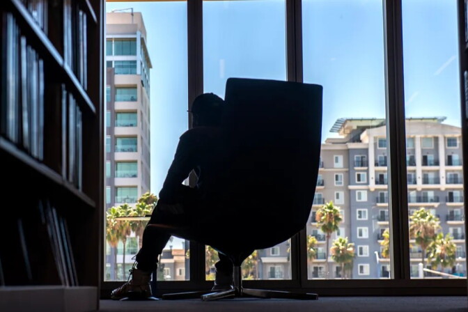 A man sits in one of the oversized chairs next to shelves of books inside a library.