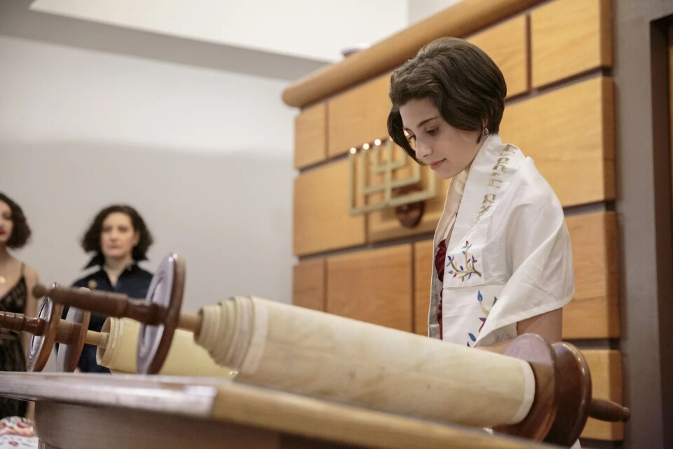 A young, white teenage girl stands in a Jewish synagogue in front of an open Torah scroll. She has short brown hair and wears a white, embroidered prayer shawl. 