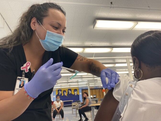A health worker in medical scrubs wearing a blue surgical mask and purple gloves holds a syringe and rolls up the sleeve of a patient, facing away from the camera. The skin of the patient's arm is exposed with the syringe hovering over it as the worker prepares to deliver a shot.