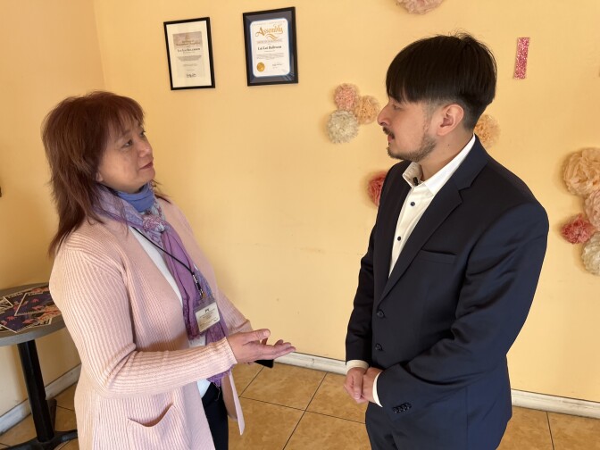 An Asian American woman in a pink cardigan and purple scarf speaks to an Asian American man in a dark suit.
