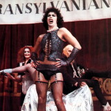 Tim Curry as Dr. Frank-N-Furter in The Rocky Horror Picture Show. He wears curly black hair, a white pearl necklace, a sparkly black vest, black panties, black gloves, and black thigh-high stockings.