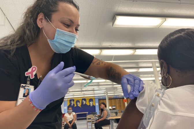 A health worker in medical scrubs wearing a blue surgical mask and purple gloves holds a syringe and rolls up the sleeve of a patient, facing away from the camera. The skin of the patient's arm is exposed with the syringe hovering over it as the worker prepares to deliver a shot.