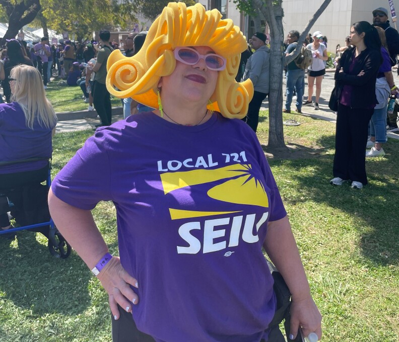 A woman wearing a plastic blonde wig, sunglasses and a purple shirt that says "SEIU" stands with a hand on her hip.