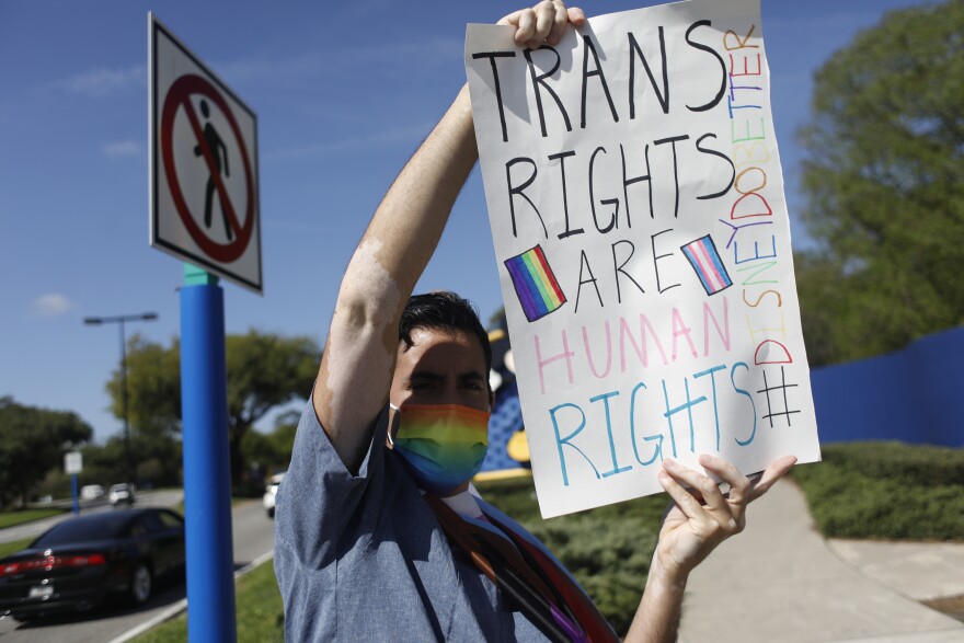 A person in a rainbow-colored mask stands on a sidewalk near a street and holds up a sign that reads: "Trans rights are human rights."
