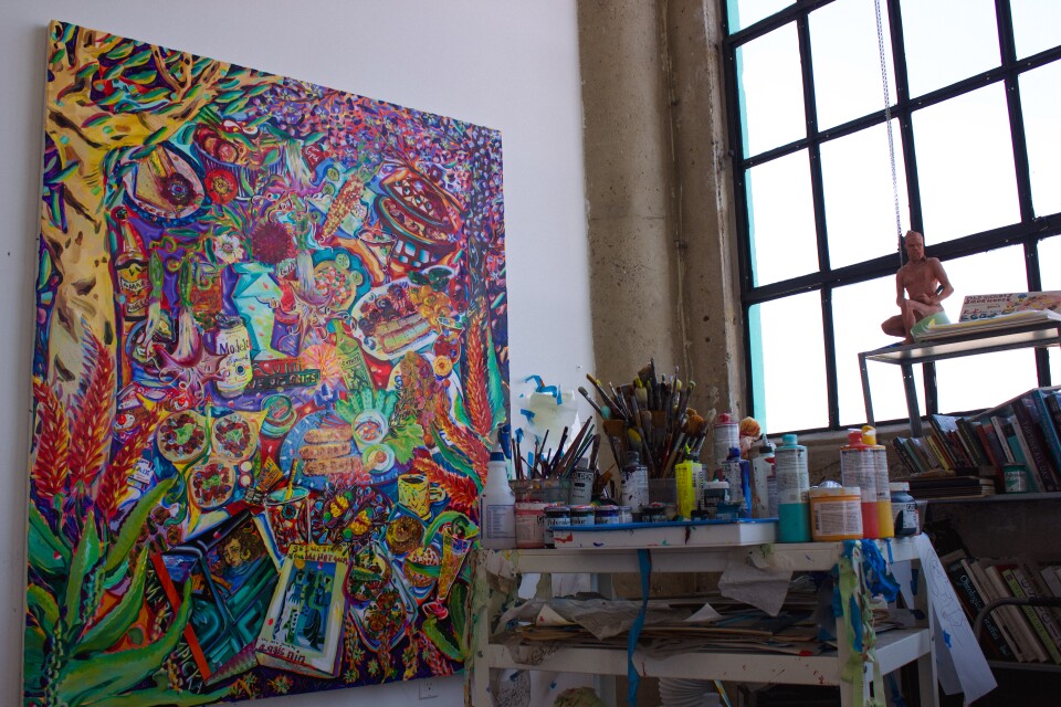 A vibrant painting on the left of the image, on the right is a cart of art supplies like paint and brushes and a cart of books.
