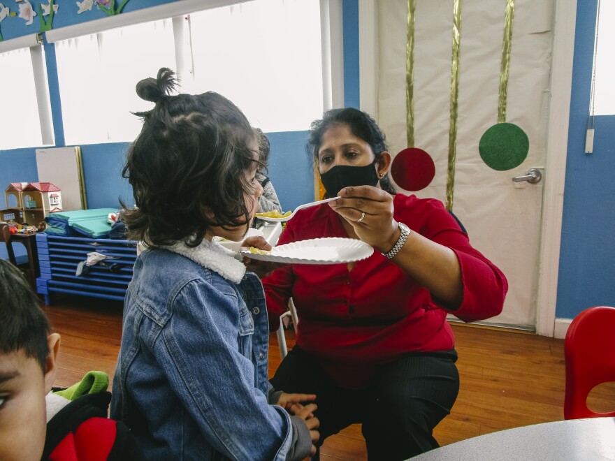 A teacher wearing red holds a plate and a utensil and feeds a child wearing a denim jacket in a classroom.