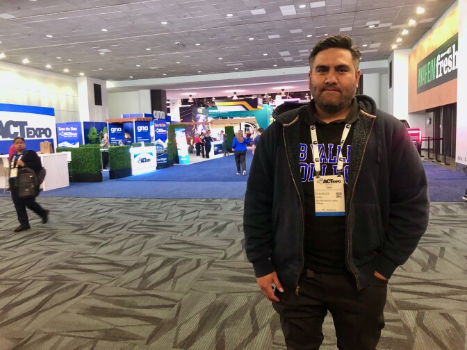 A man with light brown skin wearing a gray hoodie and black shirt stands for a photo on the carpeted showfloor of a well-lit convention center room. 