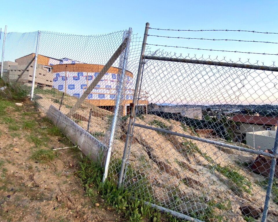 An unfinished home on the hillside behind a fence. It's a structure with exposed plywood.