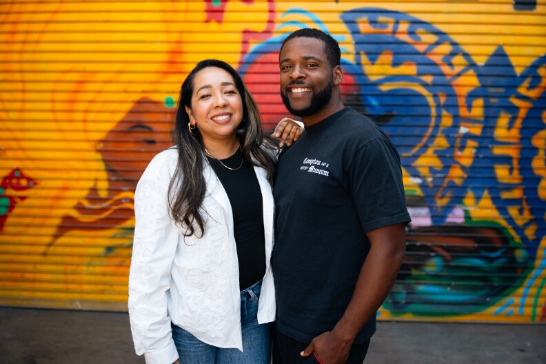 A person with dark skin-tone wearing a black T shirt and a person with medium skin-tone wearing a white jacket stand in front of a colorful blue, yellow, and brown mural.