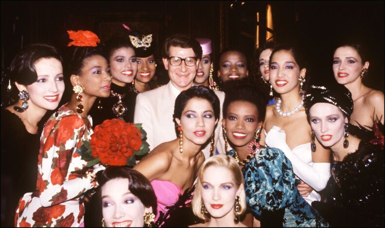 A man who appears to be white stands at the center of a group of attractive young women of all ethnicities. They are dressed very formally, with glam makeup and long earrings. They are all smiling.