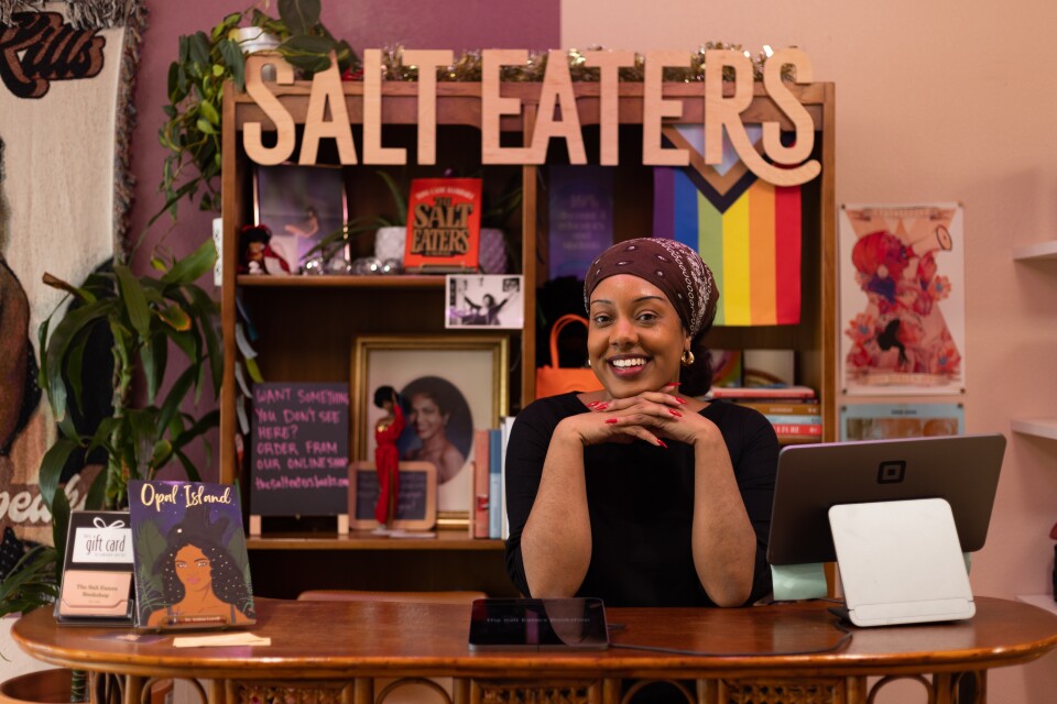 A portrait of a Black woman wearing a long sleeve black blouse, a brown bandana wrapped around her head, and red nails. She holds her hands crossed below her chin, elbows resting on a wooden table with a Square payment iPad. Behind her there's a wooden shelf with books, an LGBTQIA flag, framed photos and a wooden letters that read "Salt Eaters."