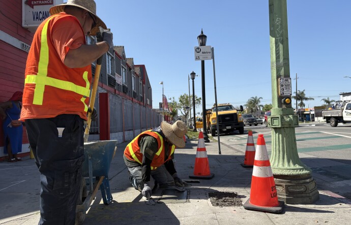 There are two men in high-visibility vests and wide-brimmed hats on a road. One is standing next to a blue wheelbarrow with a shovel. The other man is on his knees using a tool to spread wet concrete over box where street lighting wires are stored. 