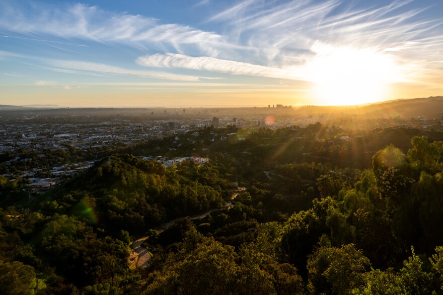 A wide view of the hills around Griffith Park, spanning down to the skyline as the sun sets with bright orange color. The hills are covered in lush green plants with dirt hiking trail visible.