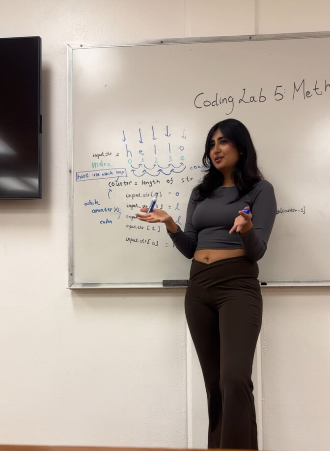 A woman, wearing black pants and a grey tshirt, stands in front of a white board gesturing with her hands. 