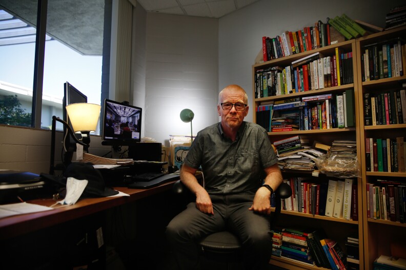 An older man wearing a chambray short-sleeve shirt and glasses sits in an office chair and poses for a photo in front of a bookcase.