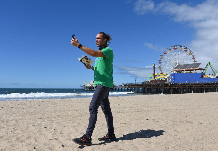 A man in a green T-shirt holding a guide book films himself doing a virtual tour of the beach in Santa Monica with the ferris wheel and pier as his backdrop.