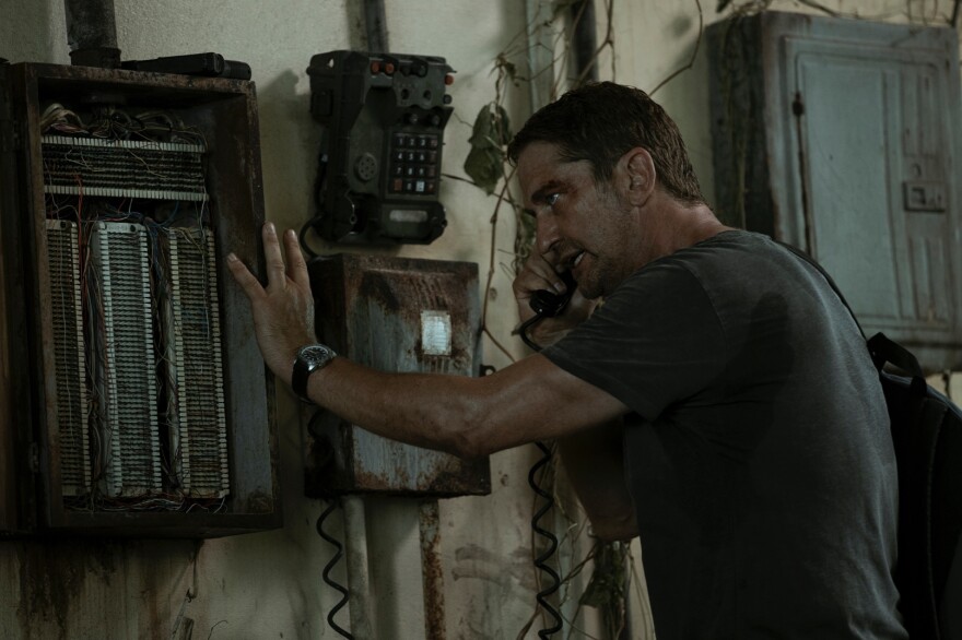 Gerard Butler stands in a dilapidated room. The white walls are stained with brown rust, a communications radio and wall phone hanging on the wall, warped and crusted with age. Butler holds the wall phone to his ear, only his left side visible as he braces himself against the wall with his left hand on the communications radio. He wears a dark gray shirt that has sweat stains. His faces is smudged with dirt and shines with sweat. His faces looks panicked and out of breath. 