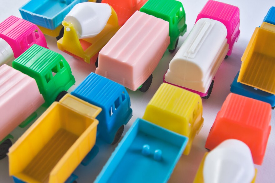 Several colorful toy trucks designed in a very simple manner, including a tank truck, a concrete transport, a refrigerator and a dump truck. Their colors are either very intense or very light. The cars are parked next to each other in two rows.