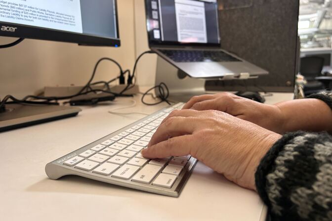 Two light skinned hands are typing on a metallic keyboard, on a desk, in front of a large screen and another laptop.