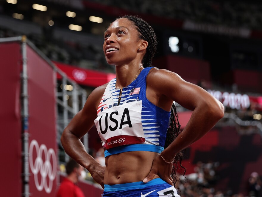 A Black woman in a racing outfit with the words U.S.A. stands with her hands on her hips smiling up to the left of the photo.