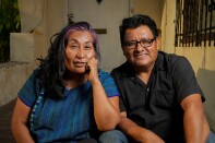 A medium-dark-skinned woman with long black and purple hair, wearing a blue embroidered blouse, and a medium-dark-skinned man wearing glasses and a black shirt sit together outside, smiling at the camera.