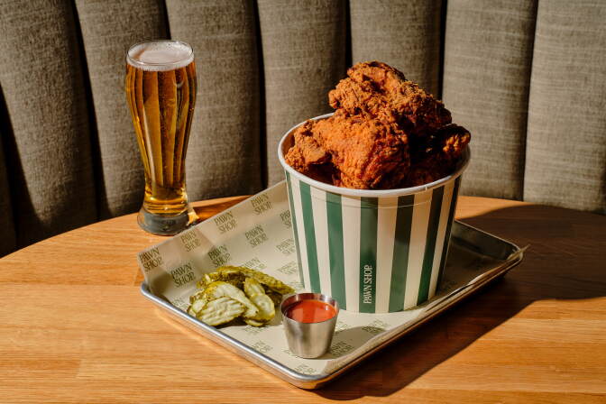 Photo of a bucket of fried chicken in metal tray, alongside it are a full sauce container, and pickles. The tray sits on a table, alongside a glass of beer.
